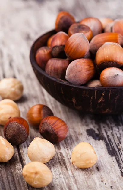 Noisettes entières en coque dans un bol en bois foncé, avec des noisettes décortiquées éparpillées sur du bois