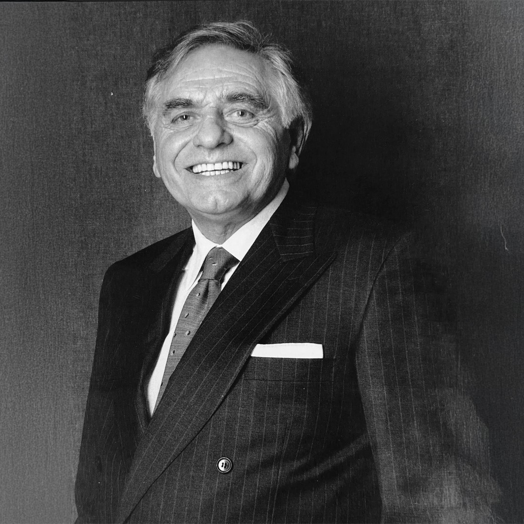 Portrait en noir et blanc d'un homme âgé souriant en costume rayé, cravate à pois et pochette, fond sombre.