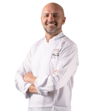 Homme chauve souriant à la barbe, bras croisés, veste de chef blanche avec logo brodé, fond uni.