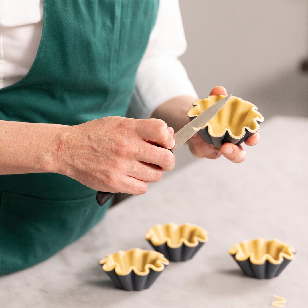 Personne en tablier vert coupe l'excédent de pâte d'un moule à tartelette noir cannelé avec un petit couteau.