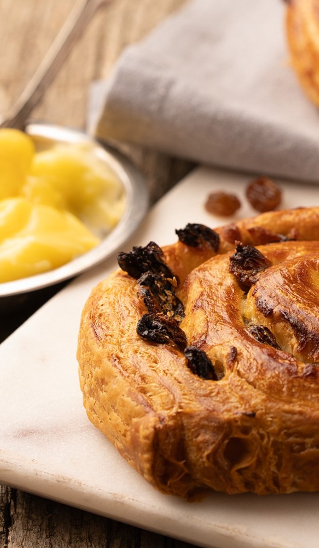 Pâtisseries en spirale aux raisins secs fourrées à la crème pâtissière sur planche blanche, raisins et bol de crème s...