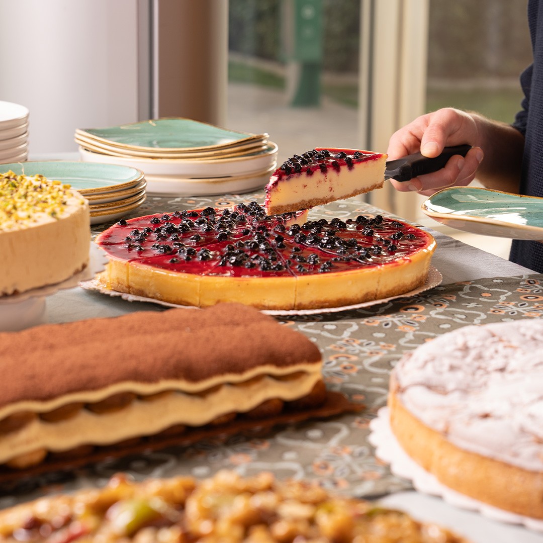 Une main sert une part de cheesecake aux myrtilles depuis une table de desserts avec tiramisu, gâteau à la pistache, ...