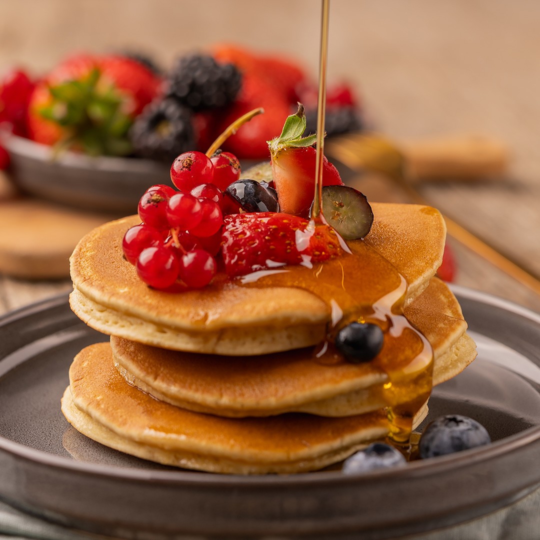 Sirop doré coulant sur des pancakes garnis de fraises, myrtilles, mûres et groseilles rouges