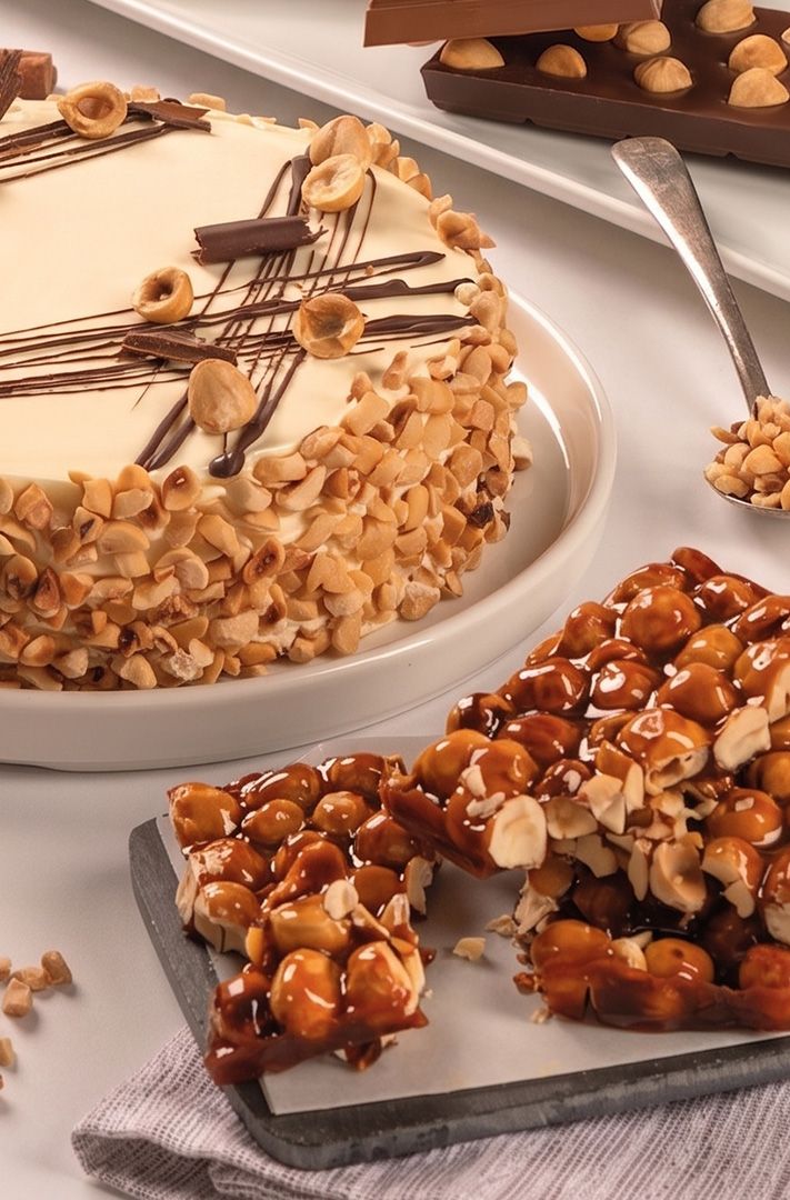 Gâteau rond garni de noisettes concassées et de filet de chocolat, à côté de carrés de croquant aux noisettes, une cu...