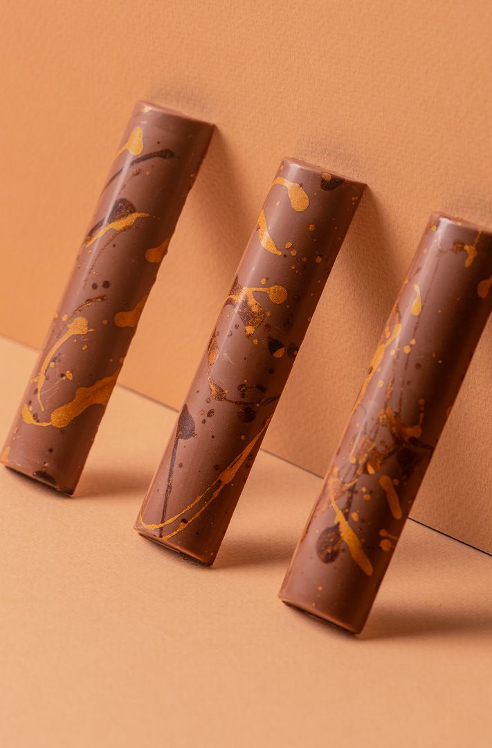 Trois tablettes de chocolat aux motifs éclaboussés dorés et foncés, appuyées contre un mur sur une surface pêche.