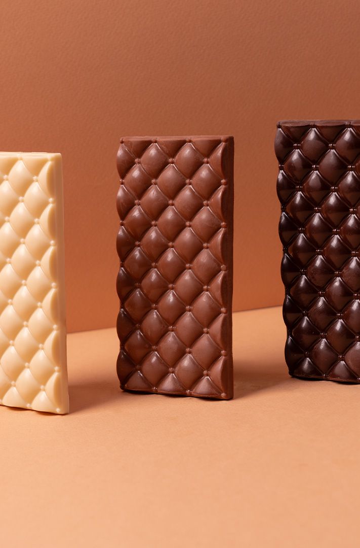 Trois tablettes de chocolat blanc, au lait et noir à texture matelassée, debout sur une surface beige