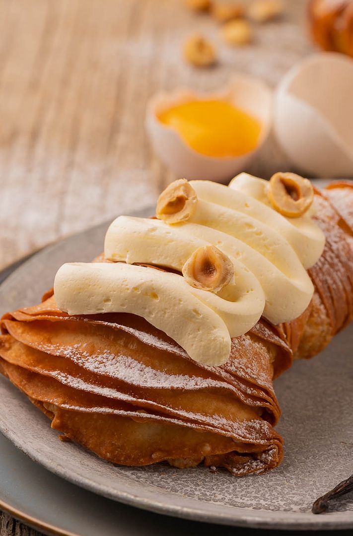 Cornet feuilleté fourré de crème et garni de noisettes sur une assiette grise, avec un œuf cassé et une gousse de van...