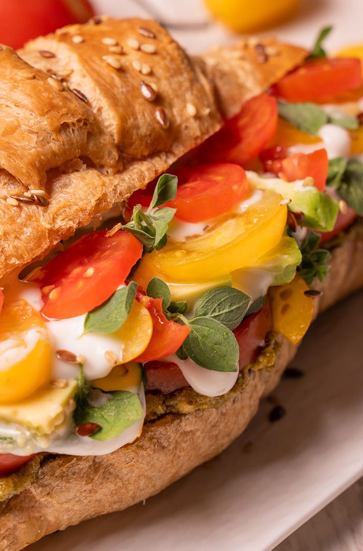 Gros plan sur un sandwich croissant aux graines, garni de tomates rouges et jaunes, avocat, herbes et sauce crémeuse