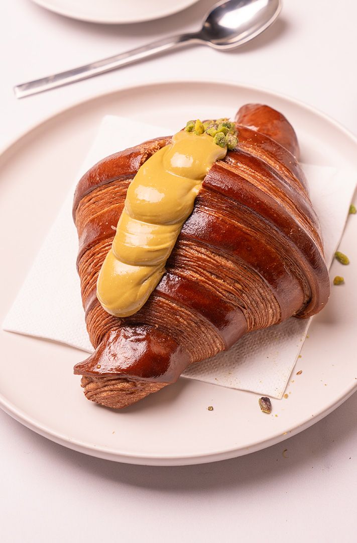 Croissant brillant garni de crème pâtissière en spirale et de pistaches concassées, servi sur assiette blanche avec c...