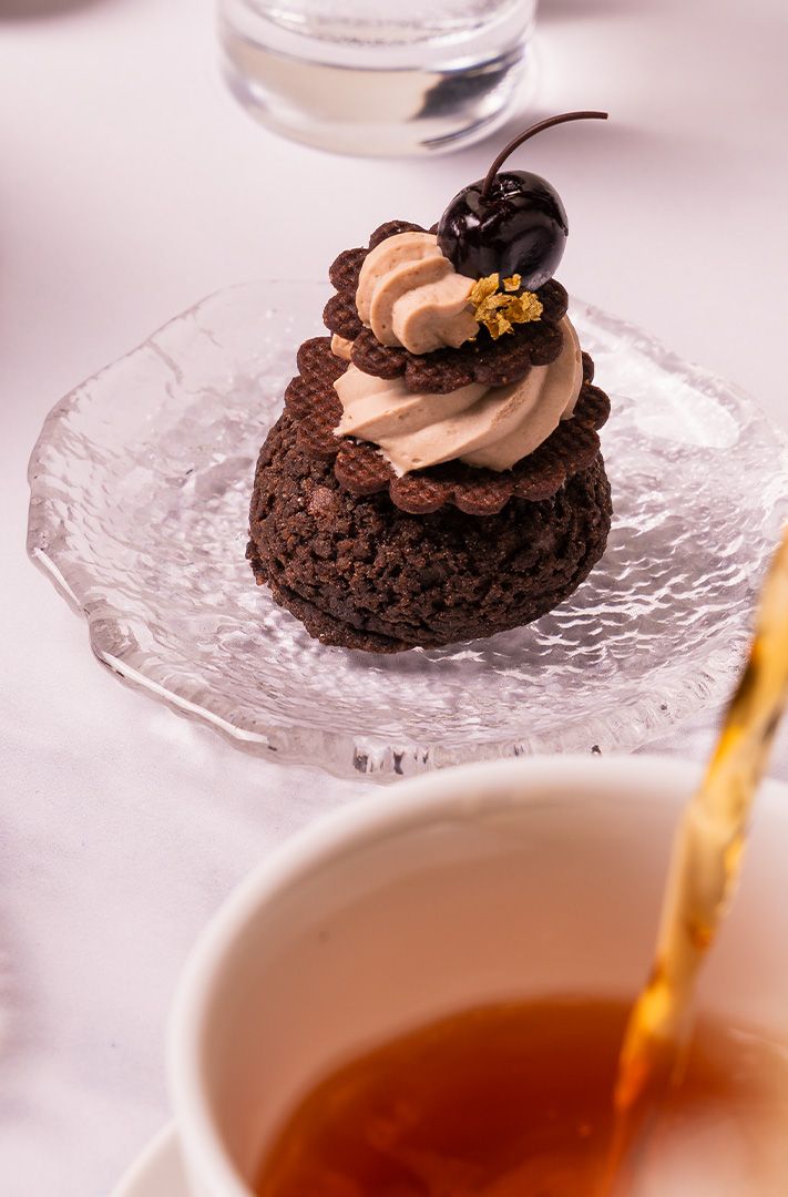Dessert de biscuit au chocolat garni de crème, cerise et feuille d'or, servi sur assiette en verre pendant qu'on vers...