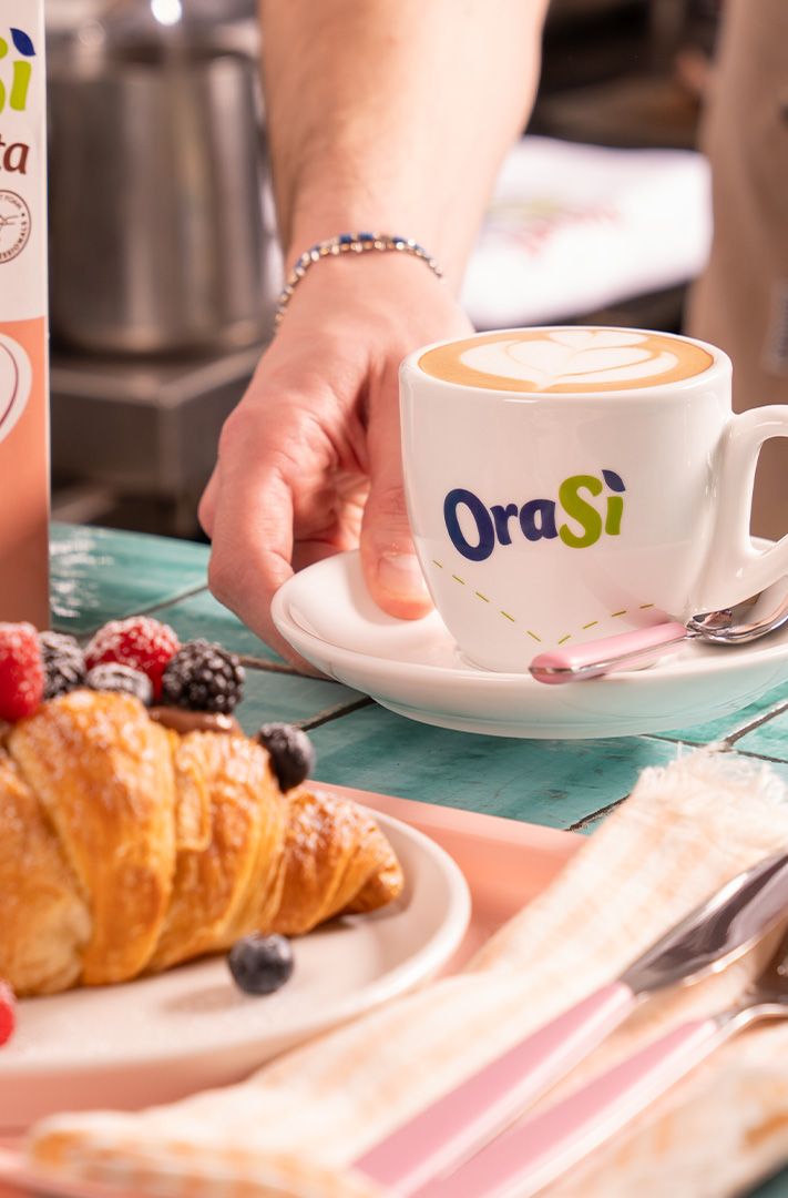 Une main pose un latte dans une tasse OraSì à côté d'un croissant aux fruits rouges et du carton de lait d'avoine Ora...