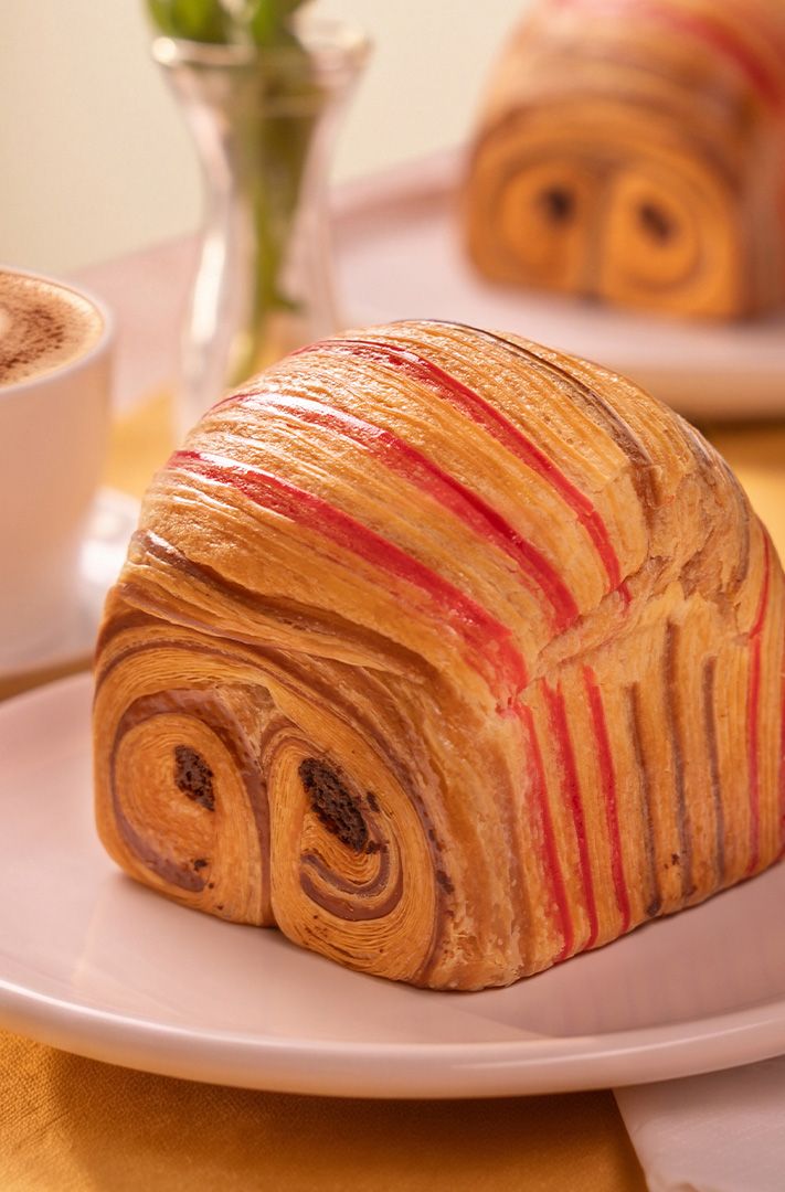 Croissant bombé à rayures rouges et motif chocolat marbré sur assiette blanche, accompagné d'un latte avec art latte.
