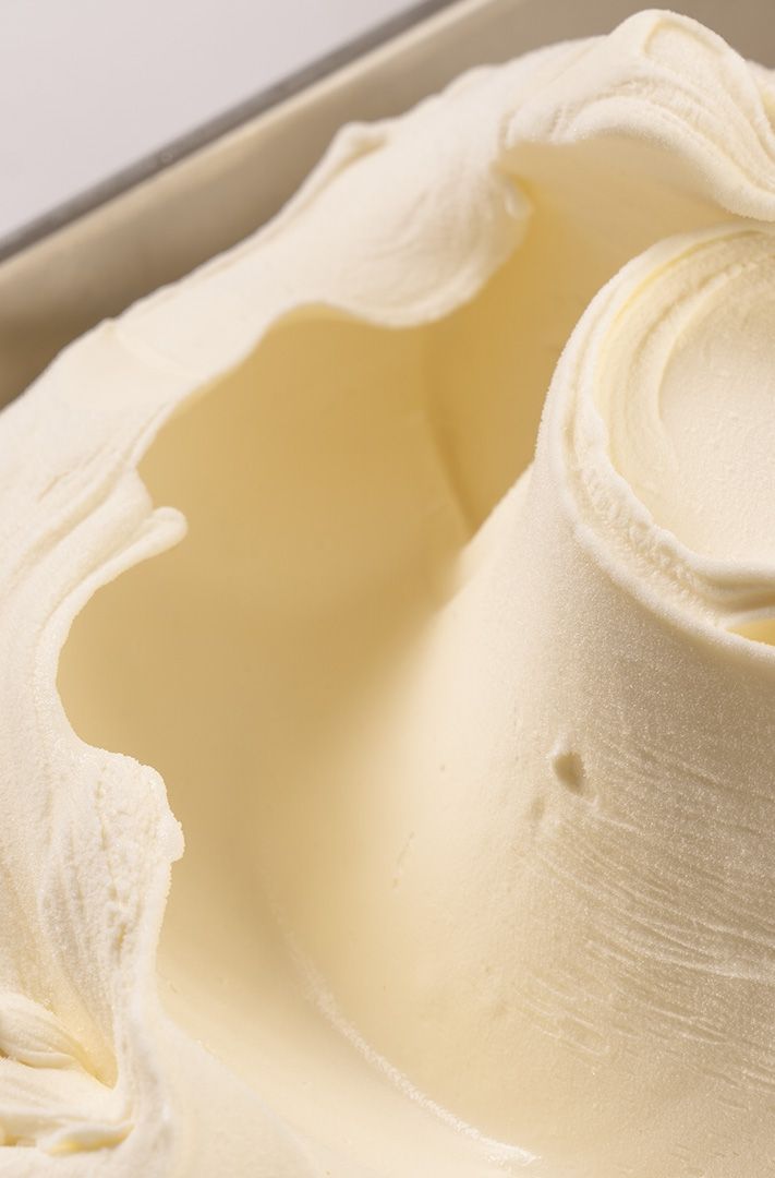 Gros plan sur une glace crémeuse jaune pâle dans un bac métallique, aux pics doux et à la texture brillante.
