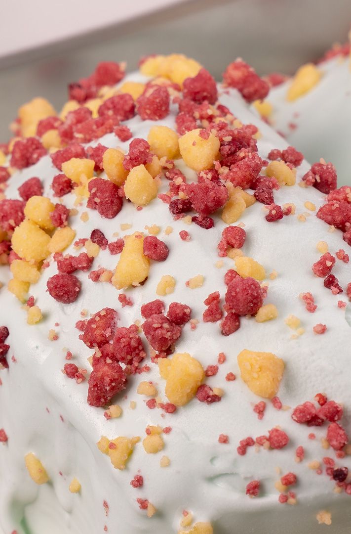 Gros plan de glace blanche parsemée d'éclats de framboise lyophilisée et de miettes jaunes.