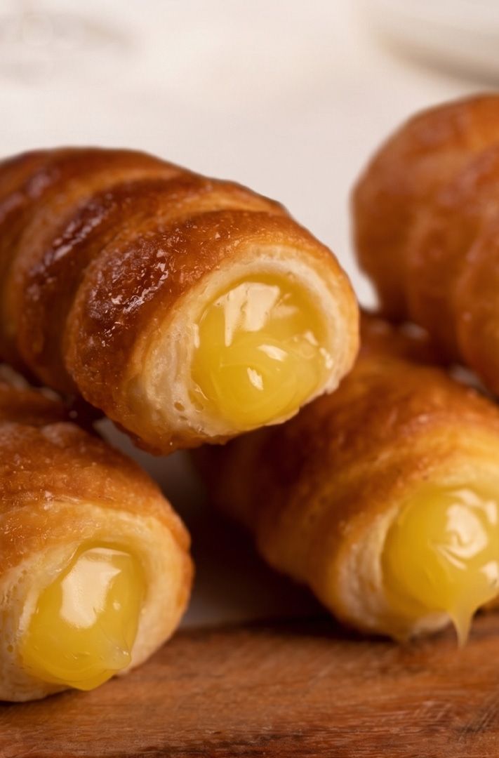 Rouleaux dorés de pâte feuilletée fourrés de crème pâtissière, empilés sur une planche en bois.
