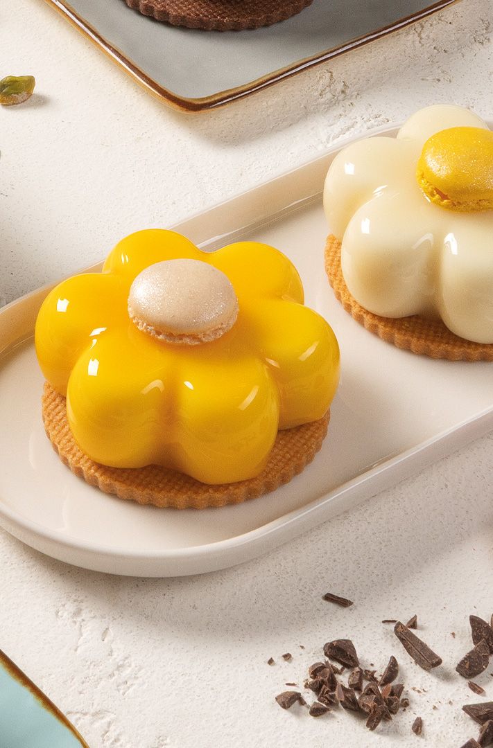 Deux tartes en forme de fleur avec glaçage jaune et blanc brillant, chacune surmontée d'un macaron, sur assiette blan...