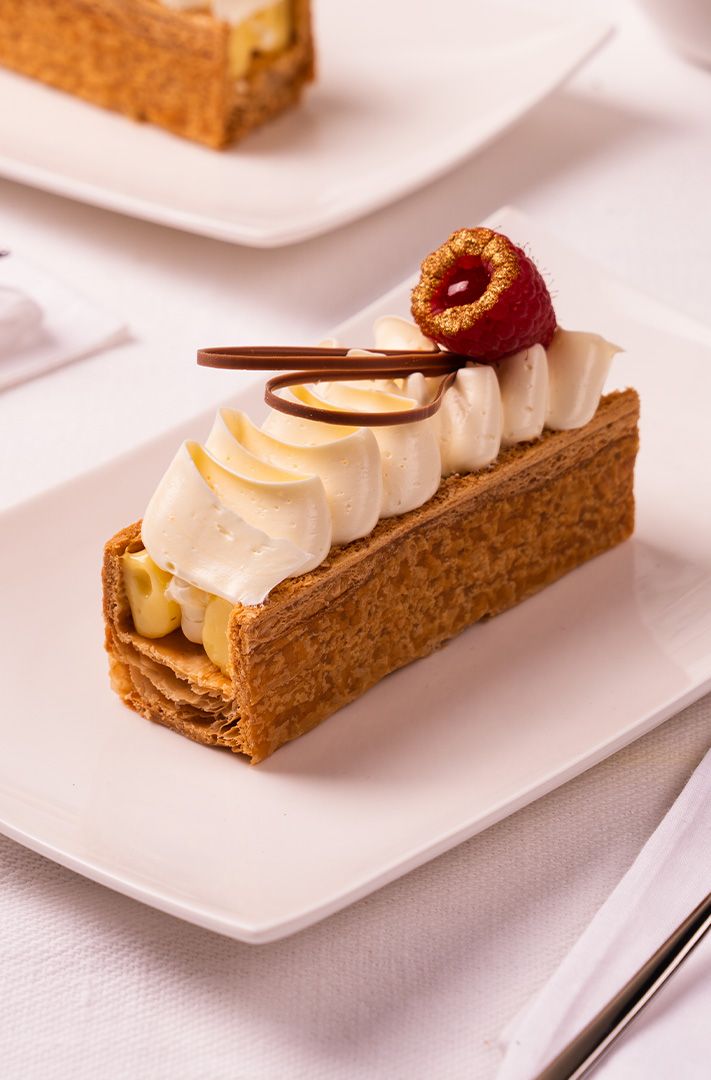 Mille-feuille garni de crème pochée, copeaux de chocolat et framboise dorée sur assiette blanche