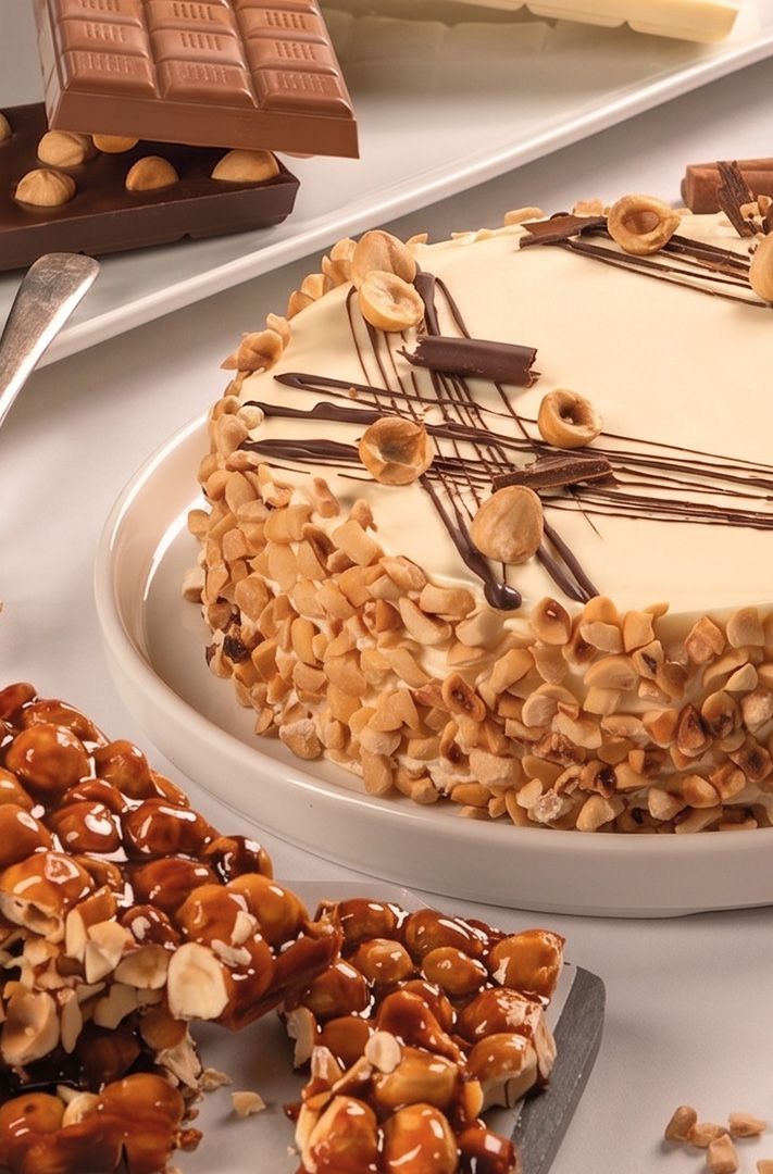 Gâteau rond à la noisette avec glaçage blanc et filet de chocolat, entouré de croquant de noisette et noisettes conca...
