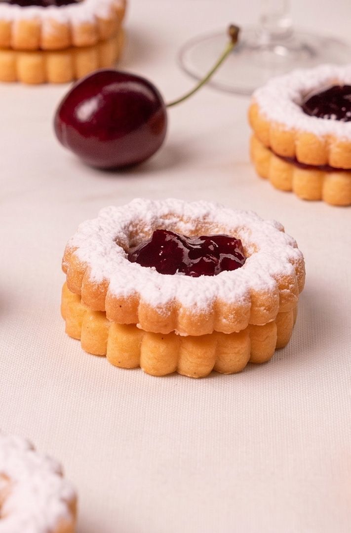 Biscuits sablés linzer festonnés, garnis de confiture rouge, saupoudrés de sucre glace et parsemés de cerises fraîches.