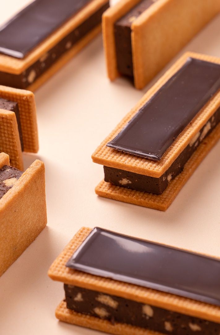 Biscuits sandwich rectangulaires fourrés au chocolat-noisette, nappés de chocolat noir brillant, sur fond crème.