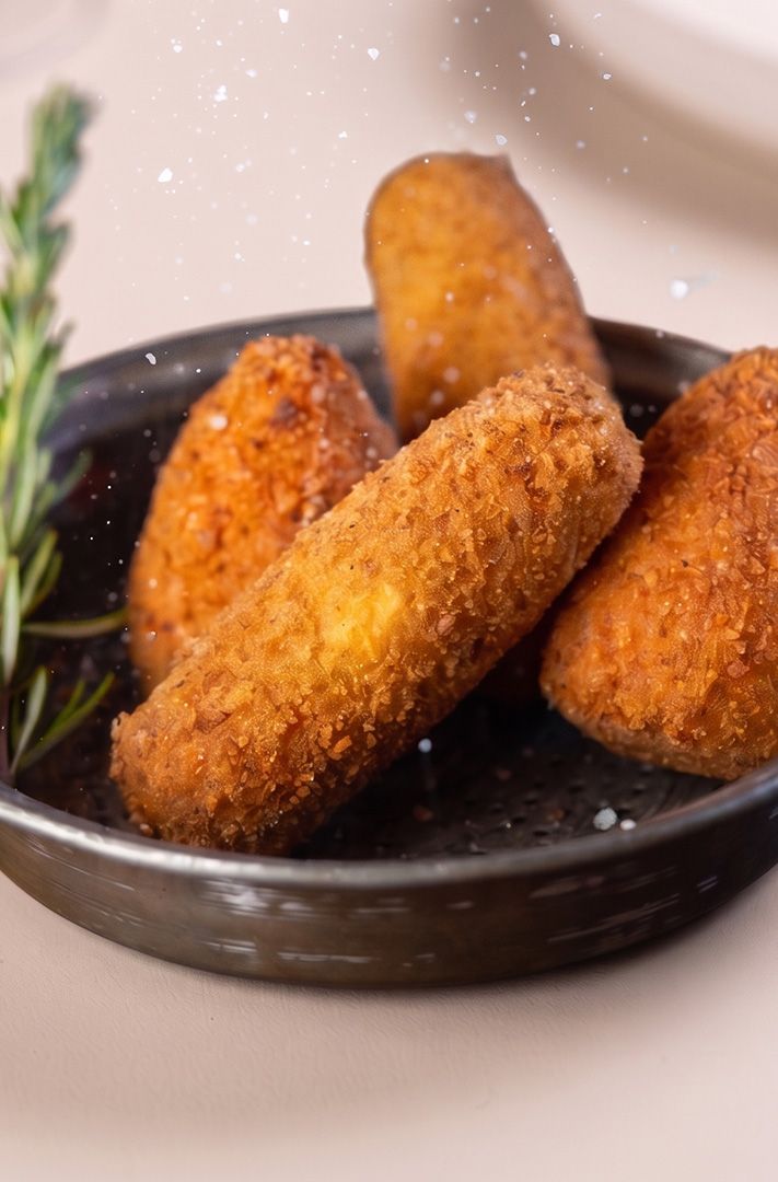 Quatre croquettes panées dorées servies dans une petite poêle sombre avec un brin de romarin frais, fond clair.